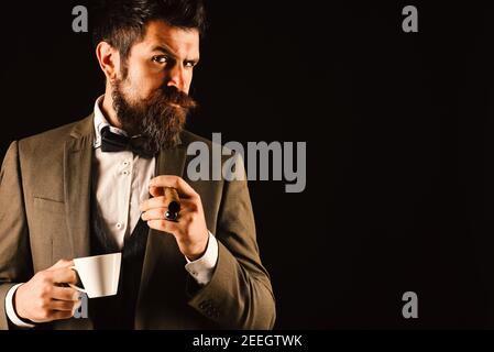 Business und schlechte Gewohnheit Konzept. Geschäftsmann mit verdächtigem Gesicht trinkt eine Tasse heißes Getränk und raucht. Mann mit Bart hält Zigarre auf schwarzem Hintergrund. Kerl in klassischen Anzug hat Morgen Kaffeepause. Stockfoto