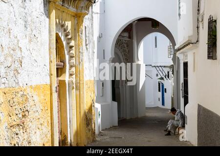 Ein Mann mit gestreifter Djellaba sitzt vor einer kleinen Moschee an der Schwelle eines Hauses in der Medina von Tanger, Marokko. Stockfoto