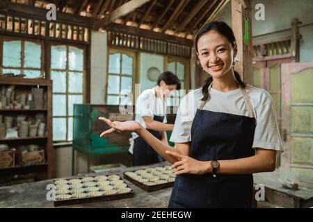 Lächelndes asiatisches Mädchen in Schürze mit Handgeste präsentieren etwas Mit Backblech-Kuchen Hintergrund auf dem Tisch Stockfoto
