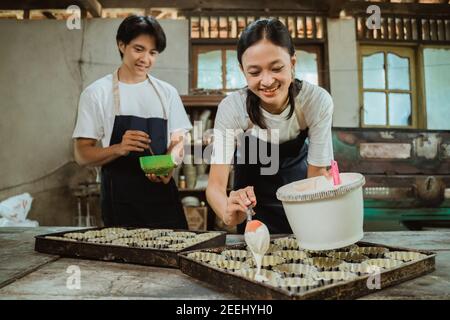 asiatische Frau lächelt trägt eine Schürze beim Gießen der Kuchen Teig auf der Kuchenform auf dem Tisch im Hintergrund Mann trägt eine Schürze hilft, den Kuchen zu backen. Stockfoto