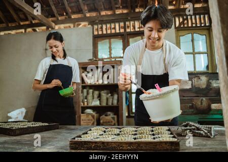 Lächelnder asiatischer Mann trägt eine Schürze beim Gießen von Teig auf einer Kuchenform auf dem Tisch im Hintergrund eine Frau trägt eine Schürze hilft, einen Kuchen zu backen. Stockfoto