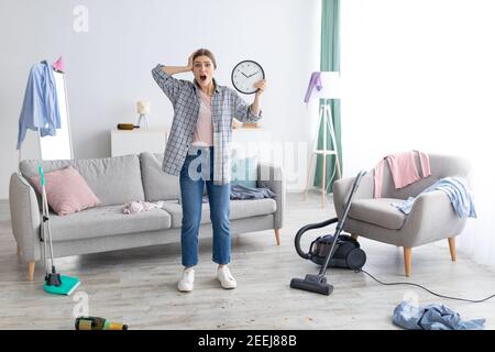 Schockierte junge Dame mit Uhr steht in einem chaotischen Zimmer nach der Party, zu spät, um Wohnung zu reinigen, bevor die Eltern zurückkehren Stockfoto