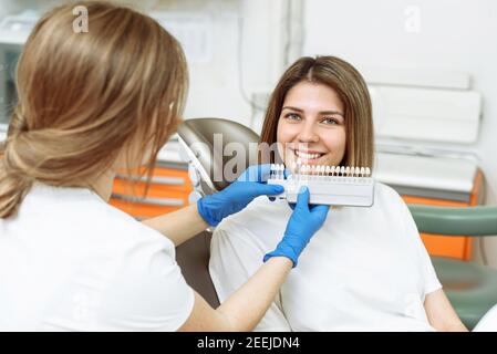 Ein Zahnarzt wählt die Farbe der Zähne einer jungen Frau zum Aufhellen aus. Zahnarzt macht den Prozess der Behandlung in der Zahnklinik Büro. Stockfoto