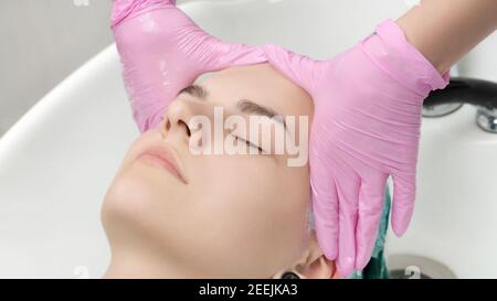 Kopf der jungen Frau mit geschlossenen Augen in Waschbecken im Friseursalon. Friseur Hände in rosa Schutzhandschuhe waschen Haar in professionellen Schönheitssalon Stockfoto