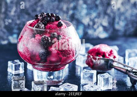 Obst- und Beereneis in einem großen schönen Glas mit Blättern aus Minze und Kugel verziert, umgeben von Eiswürfeln auf schwarzem Stein Hintergrund. Kopie, Textraum Stockfoto
