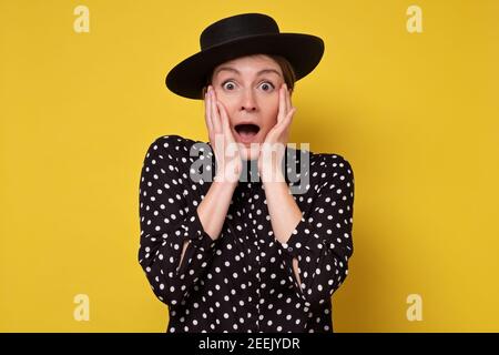 Überraschte Frau mit geöffnetem Mund und großen Augen halten Hände das Gesicht. Studio auf gelber Wand. Stockfoto