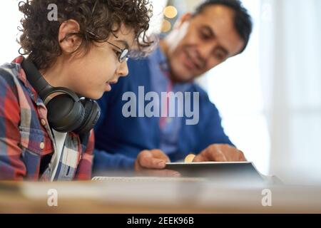 Enthusiastischer lateinischer Junge mit Brille, der konzentriert am Schreibtisch sitzt und Hausaufgaben macht, während er zu Hause Fernunterricht macht. Online-Bildung, Homeschooling Stockfoto
