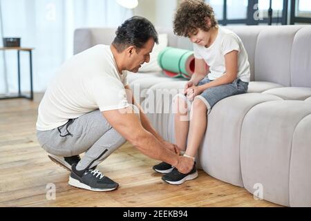 In voller Länge ein Foto von Papa, der die Schnürsenkel an einem Kinder-Sneaker bindet, während er sich für das Training zu Hause vorbereitet. Konzept der Familie Stockfoto