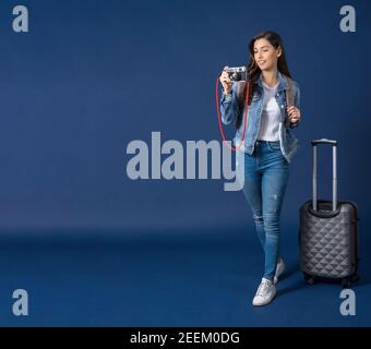 Glückliche junge asiatische Frau halten und schießen Foto mit Vintage-Kamera auf blauem Hintergrund, tragen Backpacker und Gepäck, Urlaub und Reise Konz Stockfoto