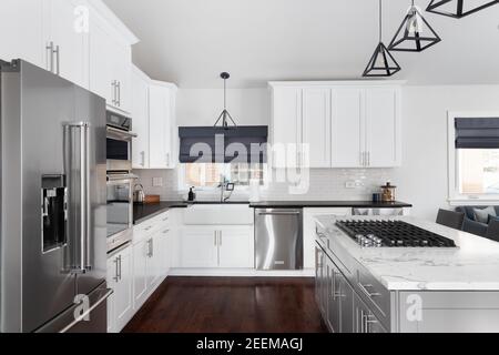 Eine wunderschöne weiße Küche in einem modernen Bauernhaus mit schwarzen Lichtern, die von der gewölbten Decke über der Insel hängen, und Barhockern. Stockfoto