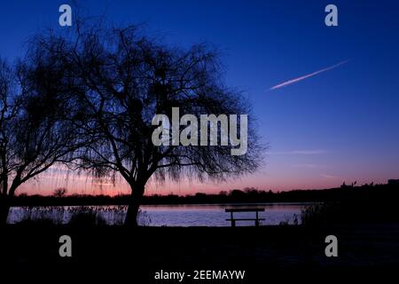 Schöner Sonnenuntergang oder Sonnenaufgang mit Blick auf einen See. Ländliche Szene mit Silhouette der Trauerweide im Vordergrund. Isolierte Bank vor dem Wasser Stockfoto