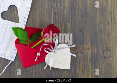 Medizinische Maske mit ausgeschnittener Herzform, roter Rose im Umschlag und Holzherzenmedaillon auf Holzhintergrund mit freiem Kopieplatz. Bleiben Sie zu Hause und qua Stockfoto