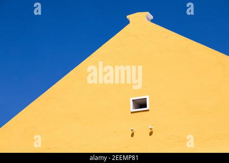 Leuchtend gelb gestrichene Giebel eines karibischen Gebäudes mit Ein kleines, weiß gerahmtes Fenster, das nach oben gegen eine klare schaut Tiefblauer Himmel mit Kopierbereich Stockfoto