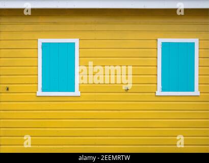 Sanft beleuchtete Seite eines gelb bemalten Holzgebäudes mit 2 geschlossenen, weiß umrahmten, blau bemalten Fenstern mit Platz für Kopien Stockfoto
