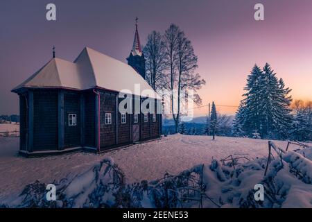 Kirche der Jungfrau Maria in Grun, Beskid-Gebirge. Schöne kleine Holzkirche. Wintermärchenbild. Der Himmel leuchtet am fernen Horizont. Stockfoto