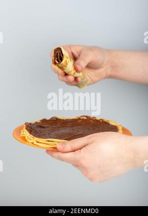 Die Hände der Frau halten einen Teller mit Pfannkuchen, der mit Schokonussbutter und einem gerollten Pfannkuchen bedeckt ist. Nahaufnahme. Stockfoto