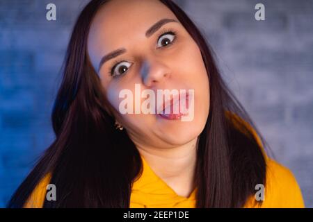 Eine junge Frau im modischen gelben Hoodie zeigt die Sprache. Porträt einer schönen Frau in einem gelben Hoodie, gegen zeigt ihre Zunge, hinten Stockfoto