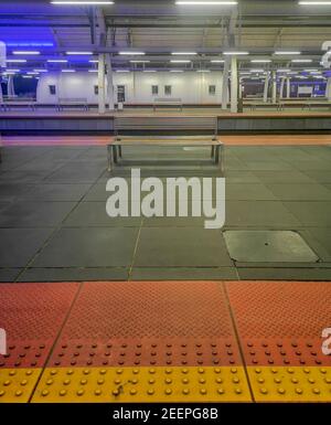 Metallbank am Bahnhof mit bunten Lichtern und rotem, gelbem und grauem Bürgersteig Stockfoto