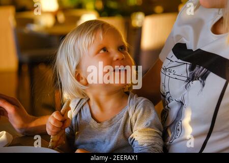 Junge blonde Mutter entspannen in sonnigen Indoor-Café Textnachrichten mit ihrem Telefon mit ihrem kleinen niedlichen Kind Pizza essen. Stockfoto