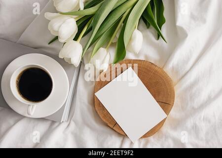 Kaffee, weiße Tulpen und leere Papierkarte auf dem Bett Stockfoto