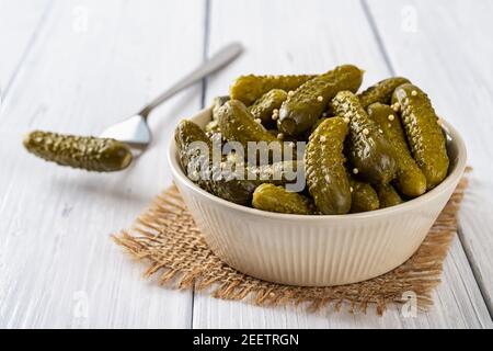 Knusprig eingelegte Cornichons in einer beigefarbenen Keramikschale und einer auf einer Gabel auf einem weißen Holztisch. Ganze grüne Gurken, mariniert mit Dill- und Senfkörnern Stockfoto