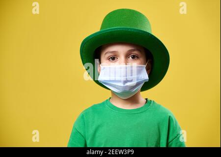 Junge in grünen irischen Kobold Hut und schützende medizinische Maske Posen zur Kamera auf einem gelben Hintergrund. Wir feiern den Tag des heiligen Patrick. Stockfoto