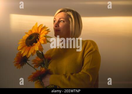 Eine Frau in einer gelben Jacke steht mit einer Sonnenblume In der Sonne im Haus auf weißem Hintergrund Und schaut aus dem Fenster.das Konzept der Hoffnung für eine E Stockfoto