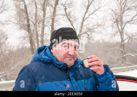 Mann mittleren Alters trinkt Tee aus Eisen Thermoskanne im Winter. Schneefall und Äste im Raureif. Ruhen Sie sich auf dem Weg in der Kälte aus. Stockfoto