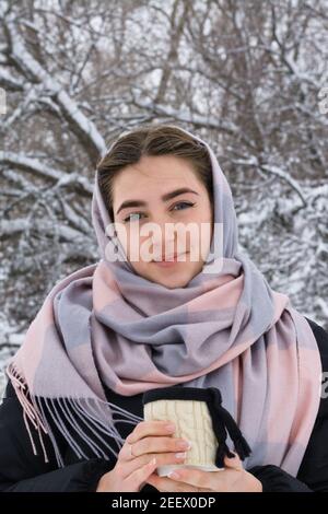 Porträt einer schönen jungen Frau mit blauen Augen. Winterspaziergang im verschneiten Wald. Becher mit heißem Getränk in der Hand. Stockfoto