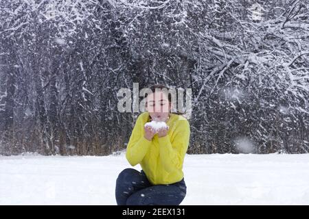 Schöne junge Frau bläst Schnee aus den Händen im Winterwald. Pulverschneemflocken fliegen hoch. Fröhliche Dame in leuchtend gelben Jacke vor Hintergrund von Whi Stockfoto