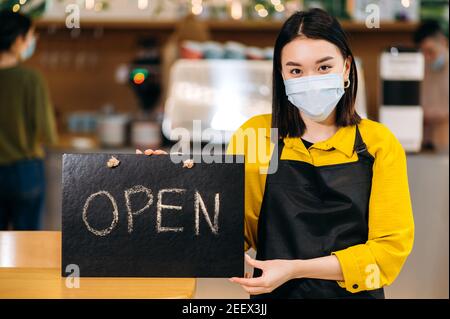 Herzlich Willkommen. Die junge asiatische Kellnerin steht im Innenbereich eines Restaurants oder Cafés mit einer schützenden medizinischen Maske und einer schwarzen Schürze und hält das Schild OFFEN. Unterstützung des Konzepts für kleine Unternehmen Stockfoto