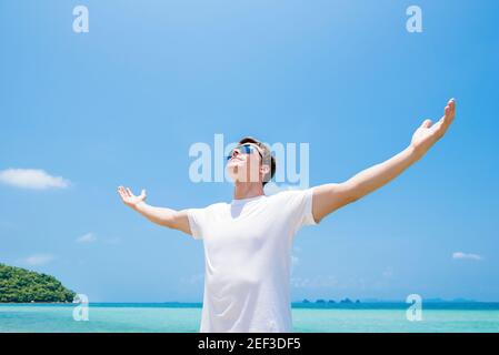 Junger Mann, der die Arme hebt, Palmen öffnet, im Sommer in die Luft am Strand blickt - Freiheit und Urlaubskonzepte Stockfoto