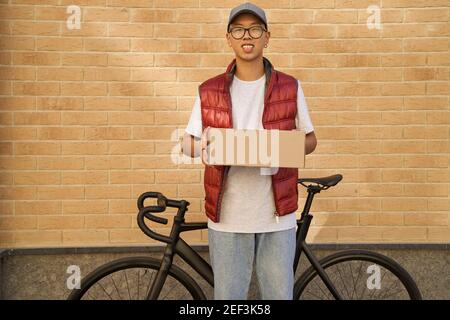 Junger asiatischer männlicher Kurier hält Paket und schaut auf Kamera, während er mit seinem Fahrrad gegen Ziegelsteinmauer steht. Konzept der Delivery Services Stockfoto