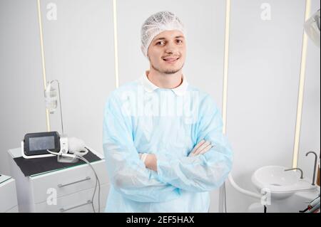 Porträt eines jungen fröhlichen Arzthelfers mit medizinischer Mütze, Mantel, stehend mit gekreuzten Armen im weißen Büro. Blick und Lächeln auf die Kamera. Zahnmedizin und Medizin Konzept. Stockfoto