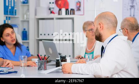 Arzt prüft die Patientenliste während des Brainstormings, diskutiert mit Kollegen am Schreibtisch. Ärzteteam spricht über Krankheitssymptome im Krankenhausbüro im Hintergrund. Stockfoto