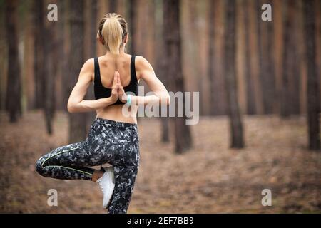 Schöne Frau, die im Wald steht und Yoga macht. Übung und Meditation Konzept. Befolgung zahlen oder Hand heben Konzept. Kiefernholz im Sommerthema. Rückansicht. Stockfoto