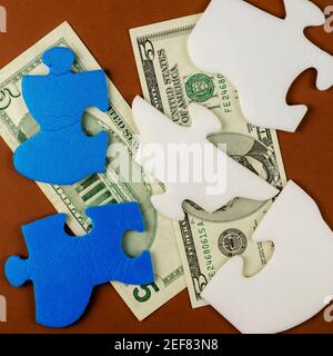 Amerikanische fünf-Dollar-Scheine sind unter den Puzzleteilen versteckt. Blau-weiße Puzzles mit Geld auf braunem Hintergrund. Draufsicht, flach liegend. Stockfoto