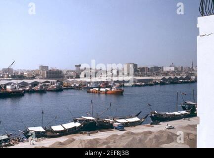 Ein Blick entlang der Uferpromenade am Dubai Creek im Jahr 1977. Der Ölreichtum des Emirats begann Ende 1960s. Es würde einen großen Unterschied machen, beginnend im Jahr 1970s, Dubai in einen globalen Staat und eine Stadt zu verwandeln. Der Golfhafen setzt immer noch auf das traditionelle Dhow (Dow), das ein- oder zweimastige arabische Segelschiff. Moderne Gebäude sind heute Teil der Landschaft. Globale Marken sind allmählich Teil der Wirtschaft – ein Zeichen für Canon ist auf einem der Gebäude im Hintergrund zu sehen. Stockfoto