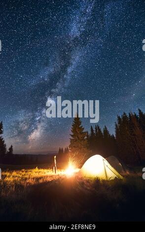 Junger männlicher Tourist, der in der Nähe eines beleuchteten Zeltes steht und unter tiefem dunklen Himmel ein hell brennendes Lagerfeuer mit vielen glänzenden Sternen und Milchstraße genießt. Camping, Tourismus im Wald, in den Bergen. Stockfoto
