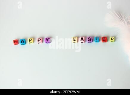 Fröhliche Ostern Schriftzug Text aus bunten Holzbuchstaben und weiße Feder auf einem weichen blauen Hintergrund. Minimalistischer Hintergrund für den Urlaub. Stockfoto