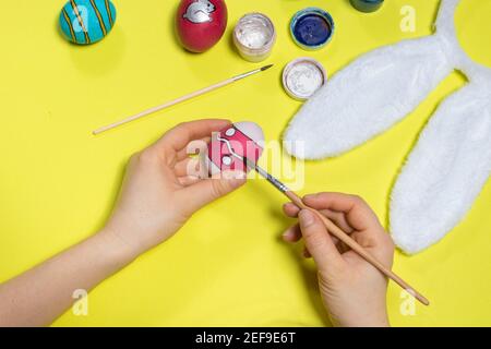 Eine Frau malt ein Hühnereier mit Gouache-Farben mit einem Pinsel auf einen gelben Hintergrund. Ostern Foto, Vorbereitung für den Urlaub. Stockfoto