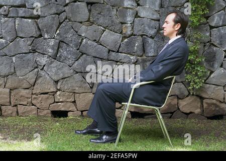 Seitenprofil eines Geschäftsmannes, der auf einem Stuhl sitzt Vorderseite einer Steinmauer Stockfoto