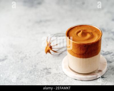 Trendiges koreanisches Getränk - Dalgona Kaffee. Glas mit kalten oder heißen Getränken - Schlagsahne Instant-Kaffee mit Milch und Schneebesen auf grauem Zement Hintergrund mit Kopierraum für Text oder Design Stockfoto