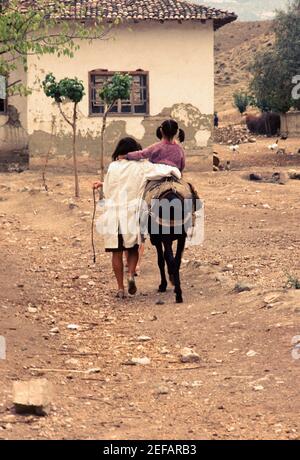 Rückansicht eines Mädchens, das auf einem Esel reitet, mit ihrer Schwester, die in einem Dorf in der Türkei entlang geht Stockfoto