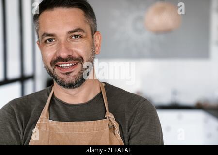 Nahaufnahme eines selbstbewussten jungen Mannes mit Schürze im Stehen Mit Küche auf einem Hintergrund drinnen Stockfoto