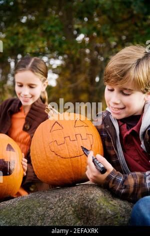 Junge, der mit seinem ein menschliches Gesicht auf einen Kürbis zeichnet Schwester, die hinter ihm sitzt Stockfoto