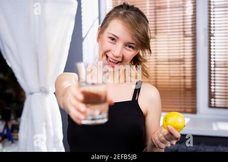 Junge Frau lächelt und hält ein Glas Wasser vor der Kamera. Innenhintergrund. Frau hält frischen Apfel . Gesundheitskonzept. Stockfoto