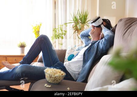 Entspannter, unterhaltsamer Mann zu Hause, der Filme in virtueller Realität sieht Gläser sitzen auf der Couch und essen Popcorn Stockfoto