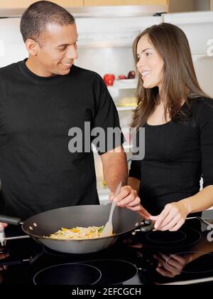 Junge Frau, die einen Mann mit mittlerem Erwachsenenalter anschaut, der Essen kocht Und lächelnd Stockfoto