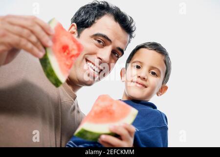 Niedrige Ansicht eines mittleren erwachsenen Mannes, der seine trägt Sohn und hält Wassermelonenscheiben Stockfoto